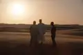 Drei Personen, zwei in traditioneller Kleidung, treffen sich in der Wüste bei Sonnenuntergang.