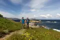 Zwei Wanderer auf einem blühenden Küstenpfad mit Blick auf das Meer.