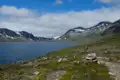 Wanderer auf einem Bergpfad neben einem See mit schneebedeckten Bergen im Hintergrund.