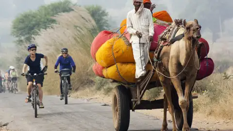Radfahrer auf einer Reise begegnen einem Kamelkarren, der mit bunten Säcken beladen ist.