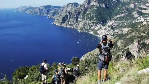 Wanderer mit Rucksäcken auf einem Bergpfad mit Blick auf die Küste und das tiefblaue Meer.