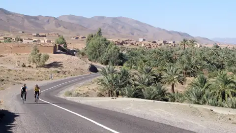 Radfahrer auf einer Straße in der Nähe einer Oase und eines Dorfes in einer bergigen Wüstenlandschaft.