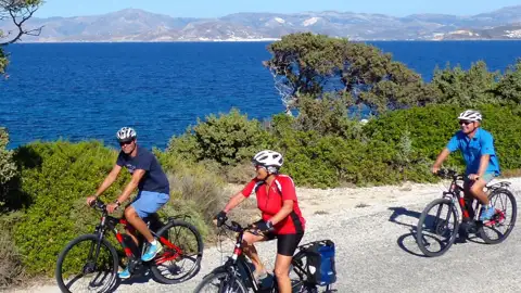Drei Personen fahren mit Fahrrädern auf einer Küstenstraße. Im Hintergrund sind das Meer und Berge zu sehen.