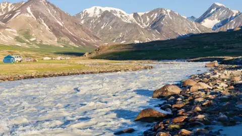 Schneebedeckte Berge, ein schnell fließender Fluss und ein Basislager im Vordergrund.