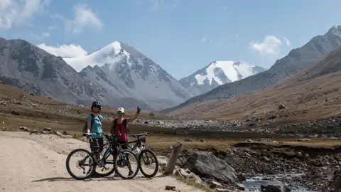 Zwei Personen auf Mountainbikes pausieren auf einem Bergpfad mit schneebedeckten Bergen im Hintergrund.