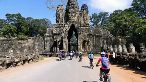 Kambodscha Radreise. Radfahrer auf einer Reise durch Angkor Thom, passieren das Südtor mit den charakteristischen Gesichtern und den steinernen Wächterfiguren.