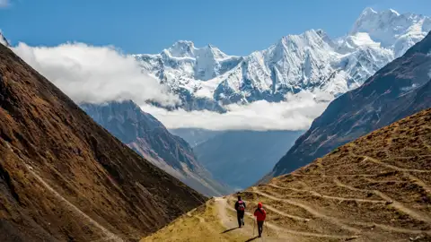 Zwei Wanderer auf einem Bergpfad im Himalaya. Im Hintergrund sind schneebedeckte Gipfel zu sehen.