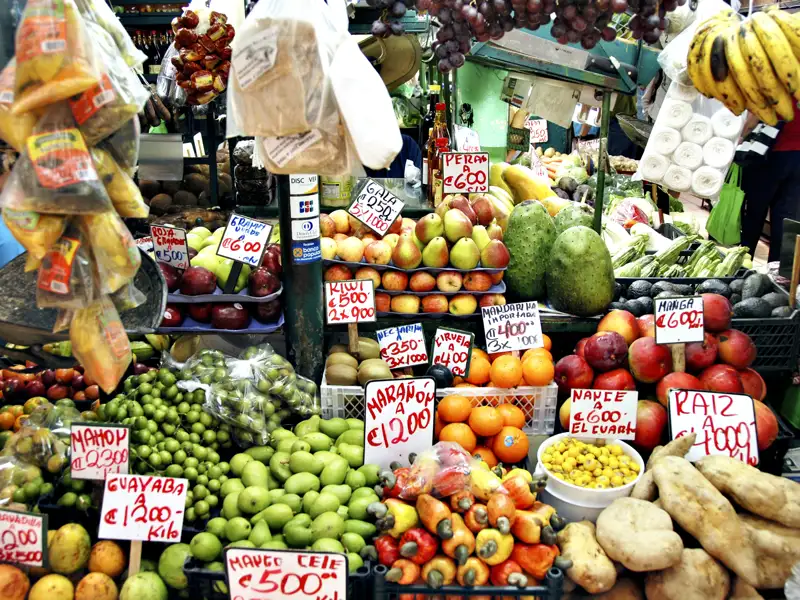 Marktstand mit tropischen Früchten und Gemüse wie Mangos, Bananen, Äpfeln, Birnen, Kiwis, Mandarinen, Guaven und Kartoffeln. Die Preise sind in Pesos angegeben.