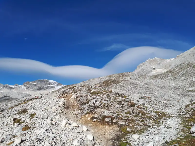 Felsiger Wanderweg in den Bergen mit markanten Wolkenformationen und schneebedeckten Gipfeln im Hintergrund.