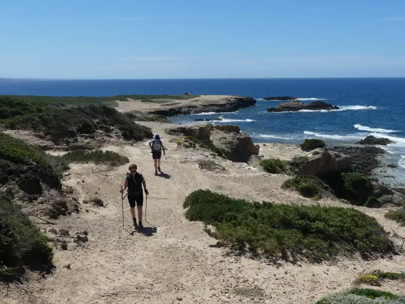 Zwei Wanderer auf einem Küstenpfad. Das Meer und felsige Küstenabschnitte sind im Hintergrund sichtbar.
