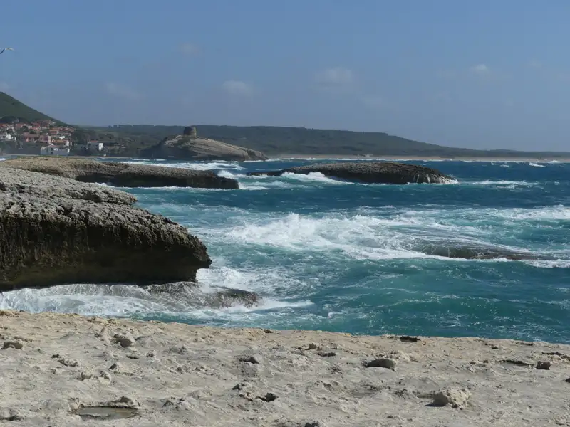 Felsige Küste mit brechenden Wellen und einem Turm in der Ferne.
