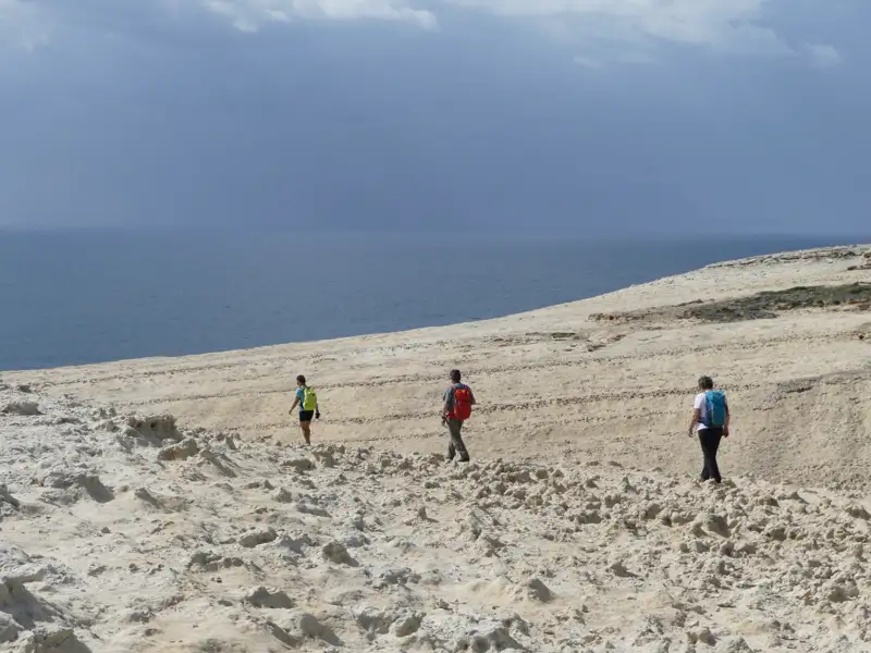 Drei Wanderer auf einem sandigen Küstenpfad. Das Meer ist im Hintergrund sichtbar.