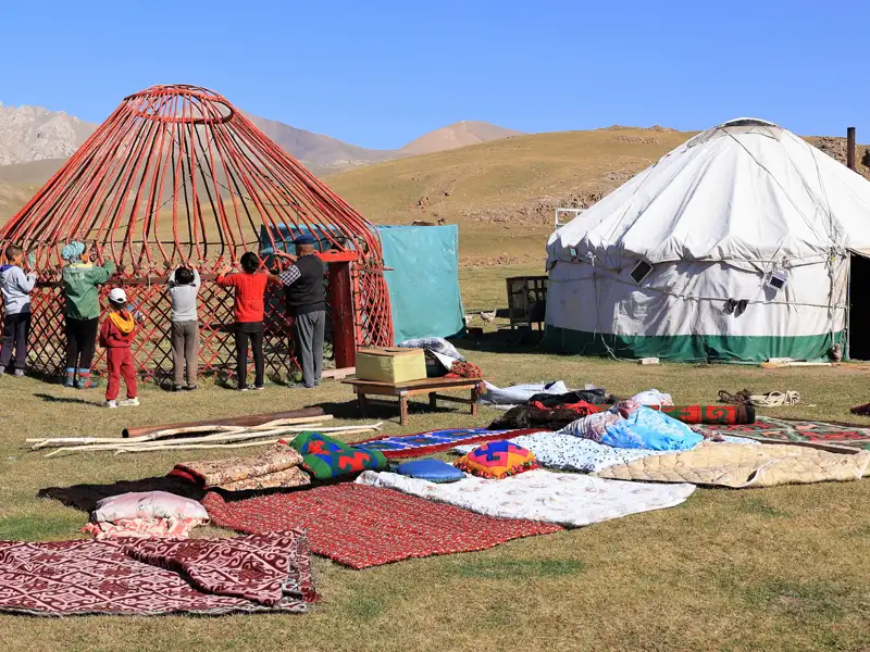 Aufbau einer Jurte in den Bergen. Mehrere Personen arbeiten zusammen, um das Gerüst der Jurte zu errichten. Im Hintergrund ist eine fertig aufgebaute Jurte zu sehen.