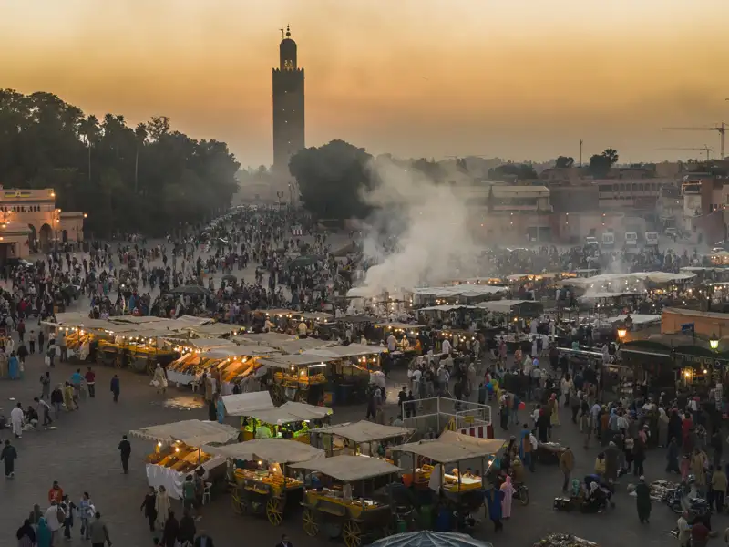 Djemaa el Fna in Marrakesch bei Sonnenuntergang mit zahlreichen Essensständen und der Koutoubia-Moschee im Hintergrund.