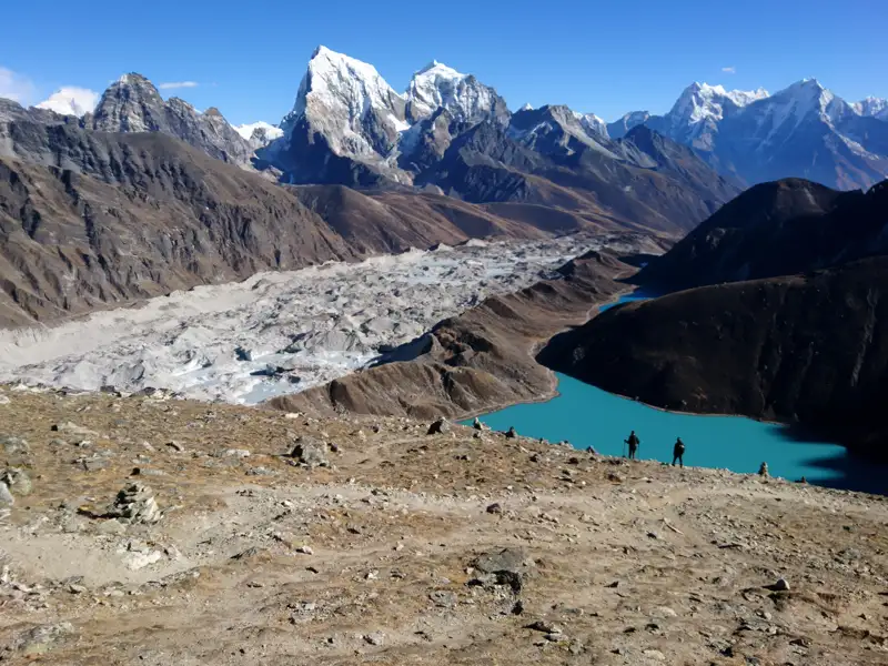 Wanderer stehen oberhalb eines türkisfarbenen Sees und eines Gletschers im Himalaya.