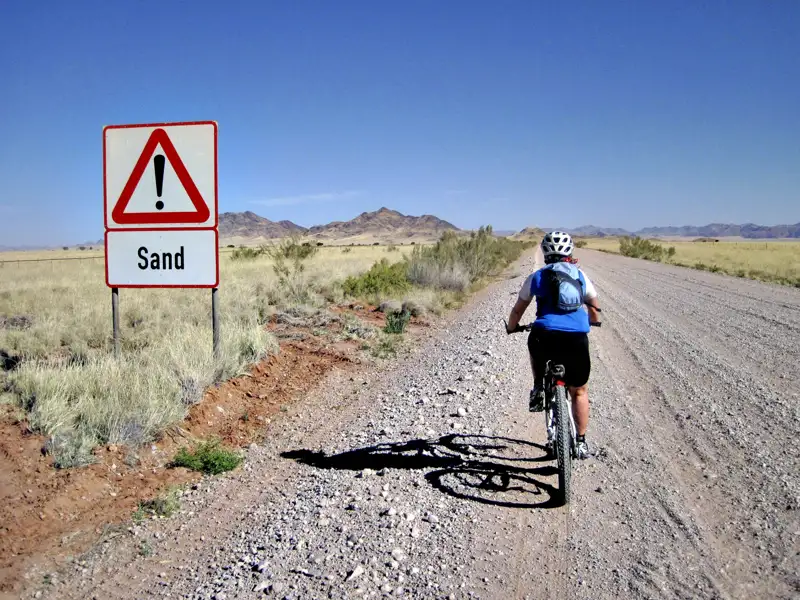 Radfahrer auf sandiger Strecke mit Warnschild.