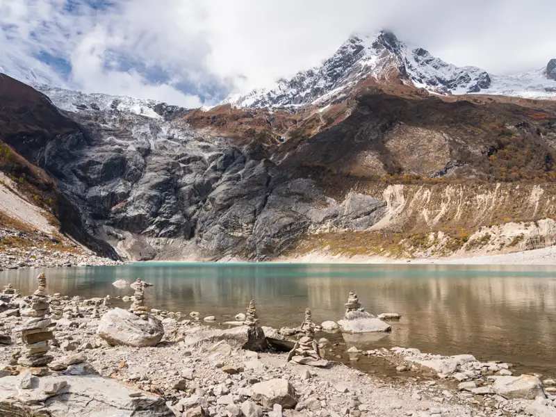 Türkisfarbener Bergsee inmitten einer Hochgebirgslandschaft mit schneebedeckten Gipfeln und Steinmännchen am Ufer.