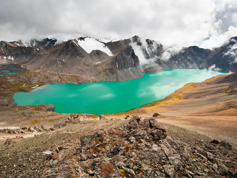 Türkisfarbener See in den Bergen mit schneebedeckten Gipfeln im Hintergrund.