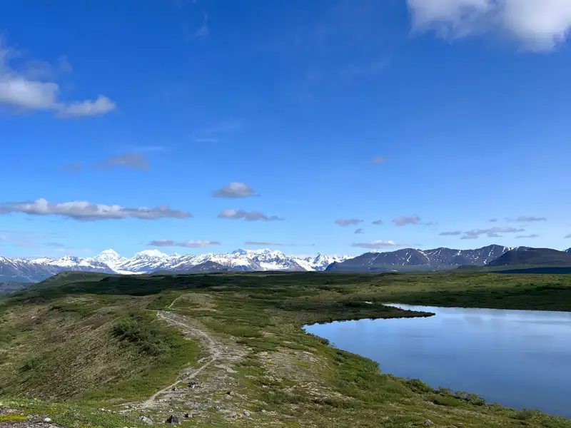 Panoramablick auf einen Wanderweg, der an einem See vorbeiführt, mit schneebedeckten Bergen im Hintergrund.