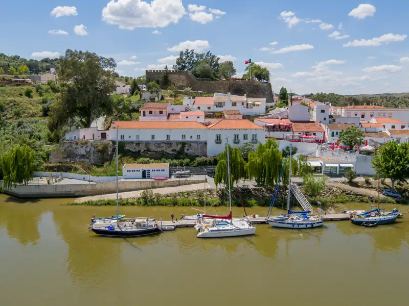 Yachthafen mit Segelbooten am Flussufer und die historische Stadt mit Festung im Hintergrund.