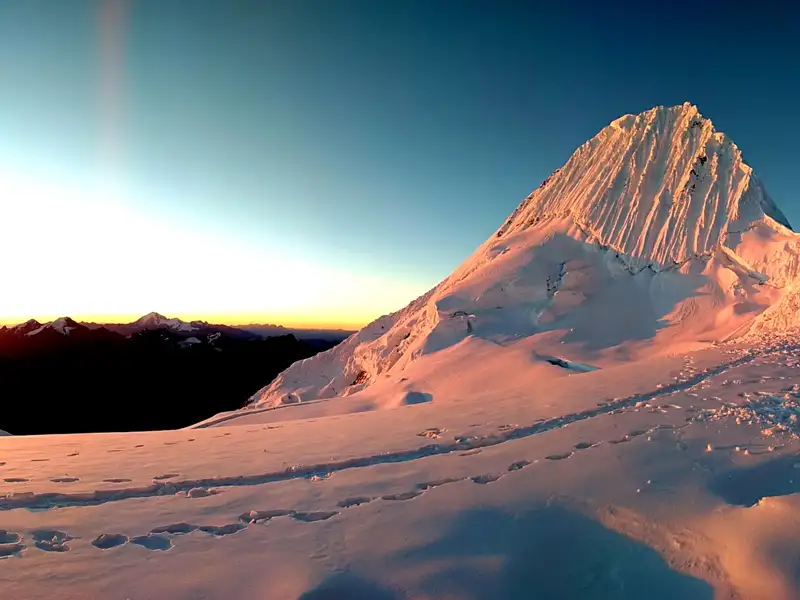 Schneebedeckter Berggipfel bei Sonnenaufgang mit Fußspuren im Vordergrund.