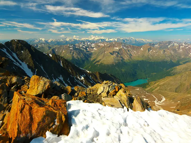 Panoramablick von einem schneebedeckten Berggipfel auf einen türkisfarbenen Bergsee und die umliegende Bergkette.