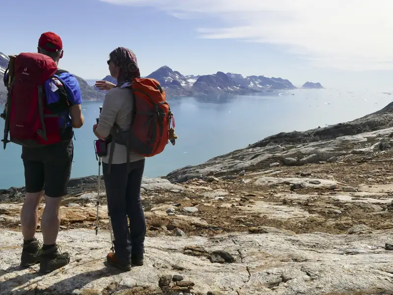 Zwei Wanderer mit Wanderstöcken und Rucksäcken stehen auf felsigem Gelände und blicken auf eine Bucht mit Bergen und Eisbergen.