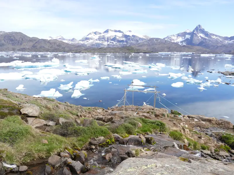 Blick auf eine Bucht mit Eisbergen und Bergen im Hintergrund. Eine Holzkonstruktion, möglicherweise für Forschungszwecke oder Fischfang, befindet sich im Vordergrund.
