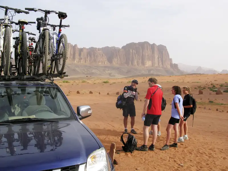 Radfahrergruppe pausiert in einer Wüstenlandschaft neben ihrem Begleitfahrzeug mit Fahrrädern auf dem Dachträger.