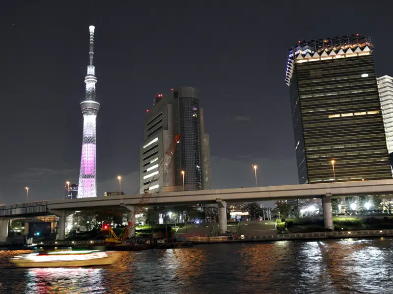 Der Tokyo Skytree bei Nacht, beleuchtet in rosa und weiß, mit umliegenden Gebäuden und einer Brücke über den Fluss.