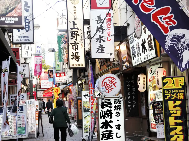 Szene einer belebten Straße in einem japanischen Marktviertel mit Schildern, die für verschiedene Restaurants, darunter "Nagasaki Suisan" (Spezialisiert auf Meeresfrüchte) und "Akebono Steak", werben.