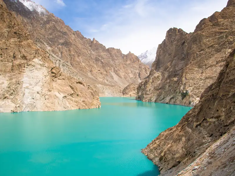 Türkisfarbener See, umgeben von felsigen Bergen mit schneebedeckten Gipfeln im Hintergrund.