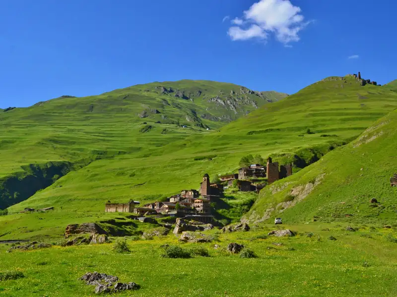 Wanderer und Packpferd auf einem Bergpfad. Im Hintergrund sind grüne Hänge und ein historisches Gebäude zu sehen.