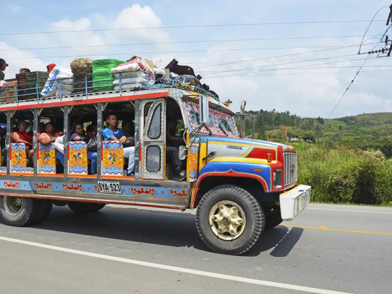 Bunter Bus mit Passagieren und Gepäck auf dem Weg durch eine ländliche Gegend.