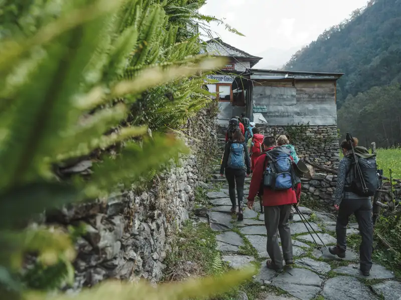 Wandergruppe auf einem Steinpfad in Richtung eines Gästehauses in einer bergigen Landschaft.