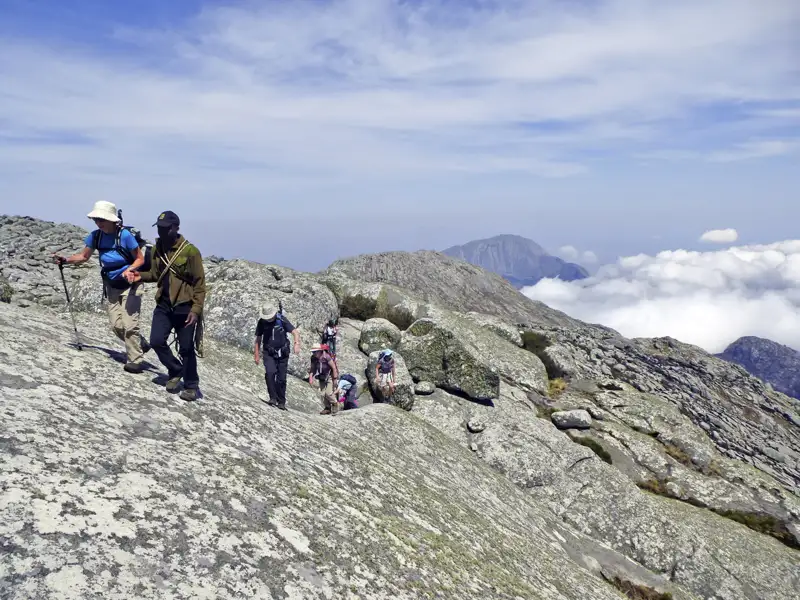Wanderer auf einem felsigen Bergpfad mit Blick auf die umliegenden Berge und Wolken.