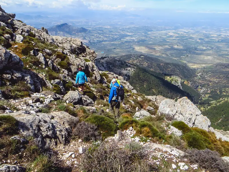 Zwei Wanderer auf einem Bergpfad, umgeben von Felsen und Vegetation, mit Blick auf ein Tal im Hintergrund.