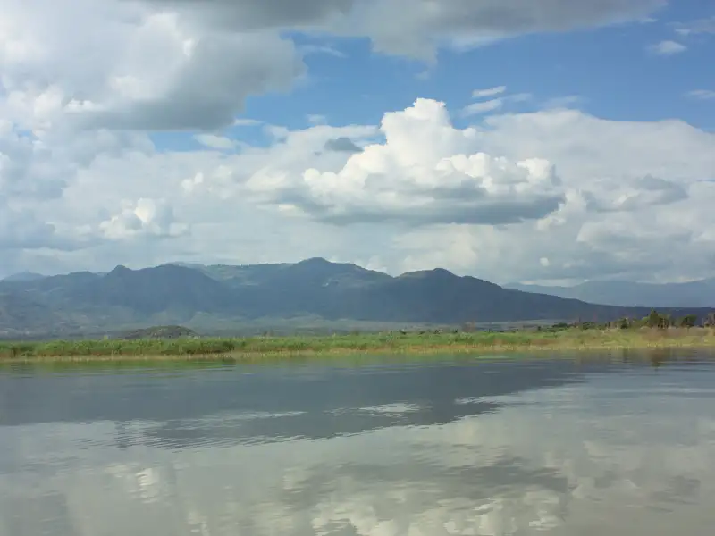 See mit Schilfgürtel und Bergen im Hintergrund unter bewölktem Himmel.