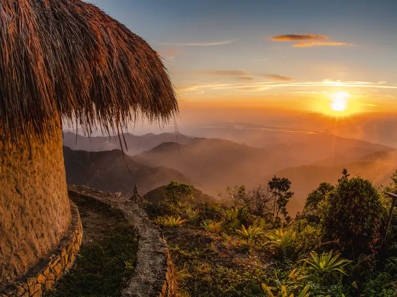 Sonnenuntergang über einer Bergkette, betrachtet von einer traditionellen Hütte mit Strohdach aus.