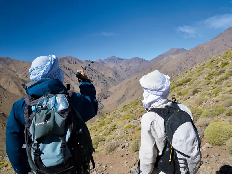 Zwei Wanderer mit Rucksäcken und Kopftüchern stehen in einer bergigen Landschaft und schauen in ein Tal.