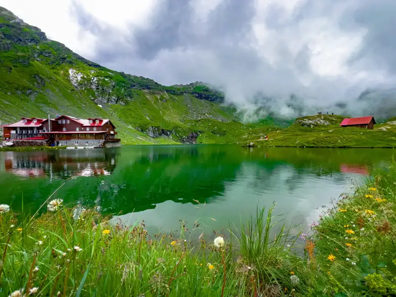 Panorama eines Bergsees mit einem roten Gebäude am Ufer und Wolkenverhangenen Bergen im Hintergrund.