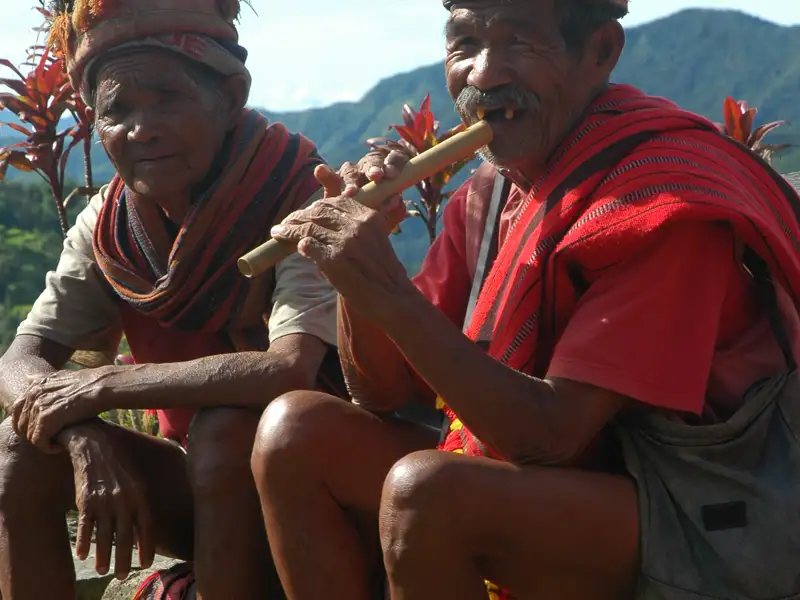 Zwei Personen in traditioneller Kleidung, möglicherweise Angehörige einer indigenen Gruppe, sitzen in einer bergigen Landschaft. Eine Person spielt auf einer traditionellen Flöte.