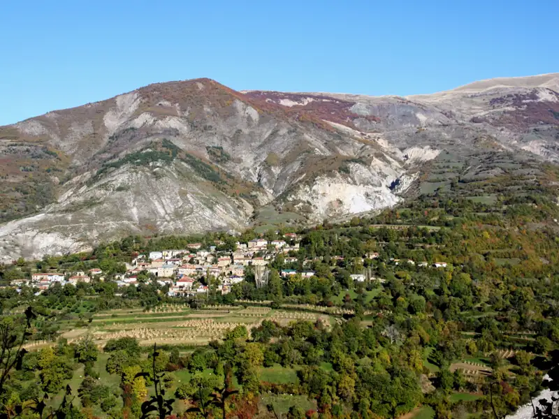 Panoramablick auf ein Bergdorf, umgeben von herbstlicher Vegetation und felsigen Berghängen.