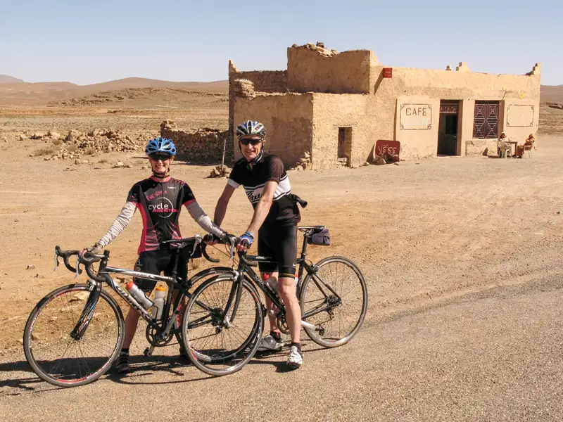 Zwei Radreisende pausieren vor einem Café in der Wüste.