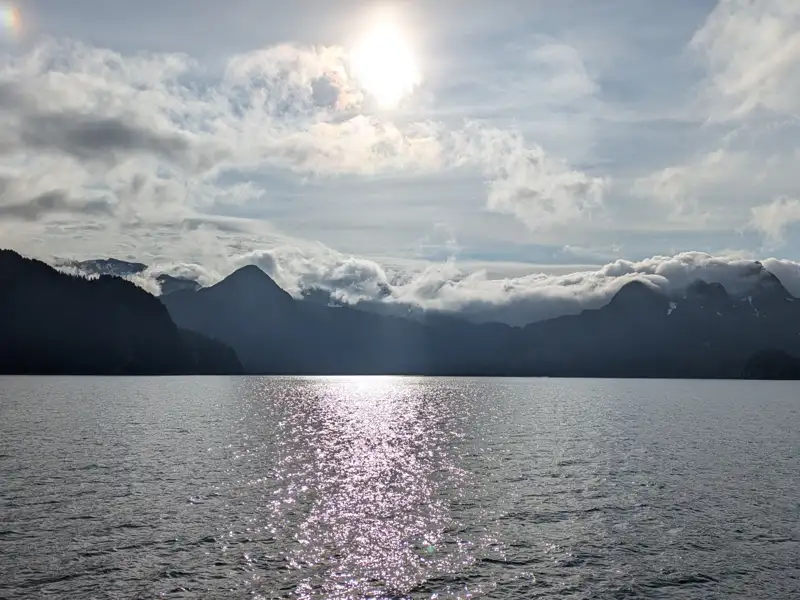 Sonnenlicht glitzert auf einem See, im Hintergrund sind bewaldete Berge und Wolkenformationen zu sehen.