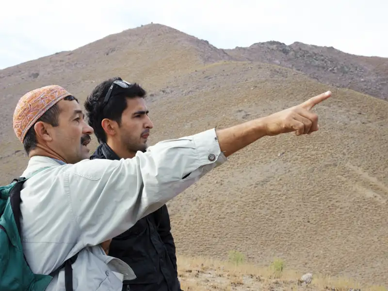 Zwei Männer in einer bergigen Landschaft, einer zeigt auf einen bestimmten Punkt.