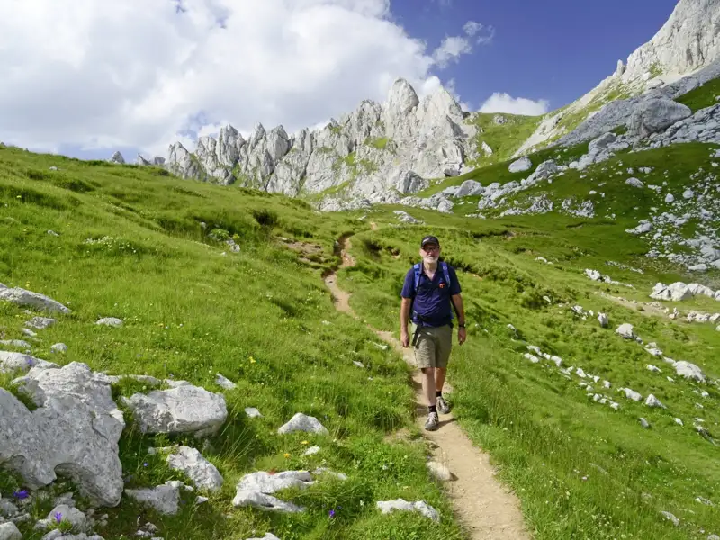 Wanderer auf einem Bergpfad mit felsigen Gipfeln im Hintergrund.