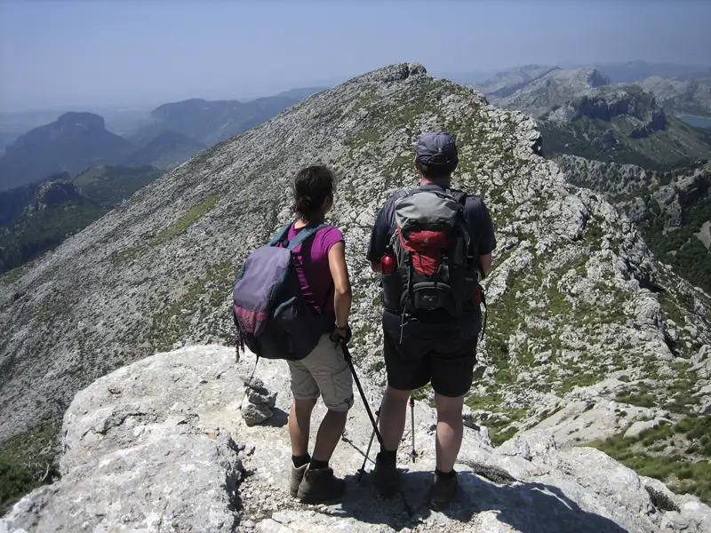 Zwei Wanderer mit Wanderstöcken stehen auf einem Berggipfel und schauen auf das Panorama.