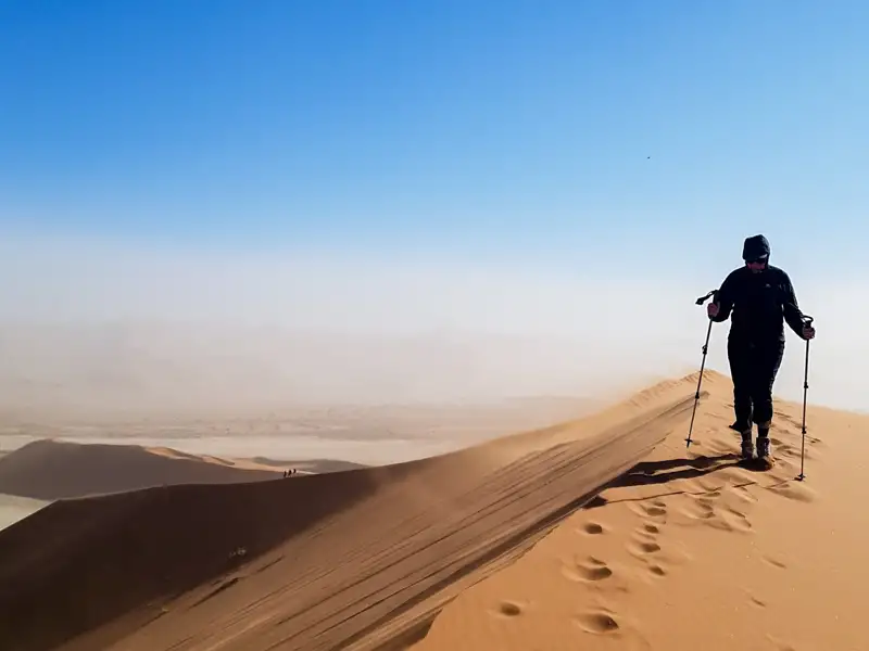 Person besteigt eine Sanddüne mit Wanderstöcken.