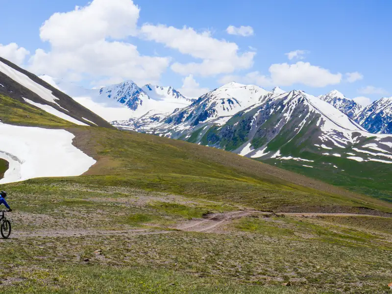 Mountainbiker auf einem Bergpfad mit Schnee und Bergen im Hintergrund.